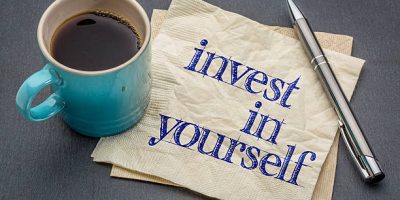 Invest in yourself advice or reminder - handwriting on a napkin with cup of coffee against gray slate stone background